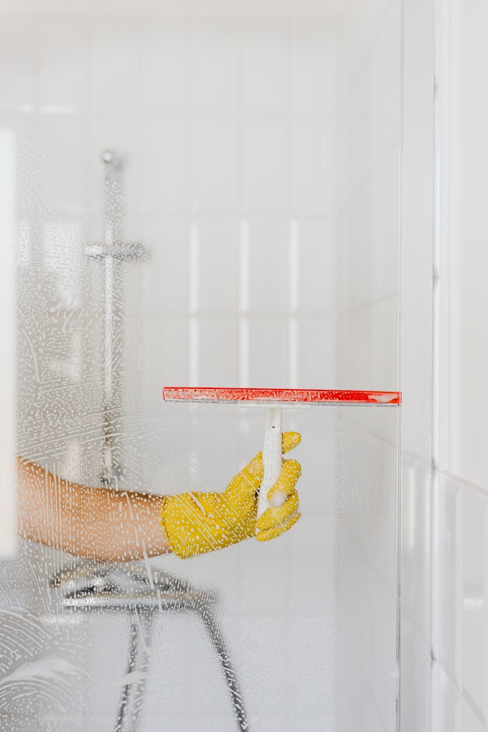 about-us A person wearing a yellow glove cleans a shower glass with a squeegee, emphasizing hygiene and cleanliness.