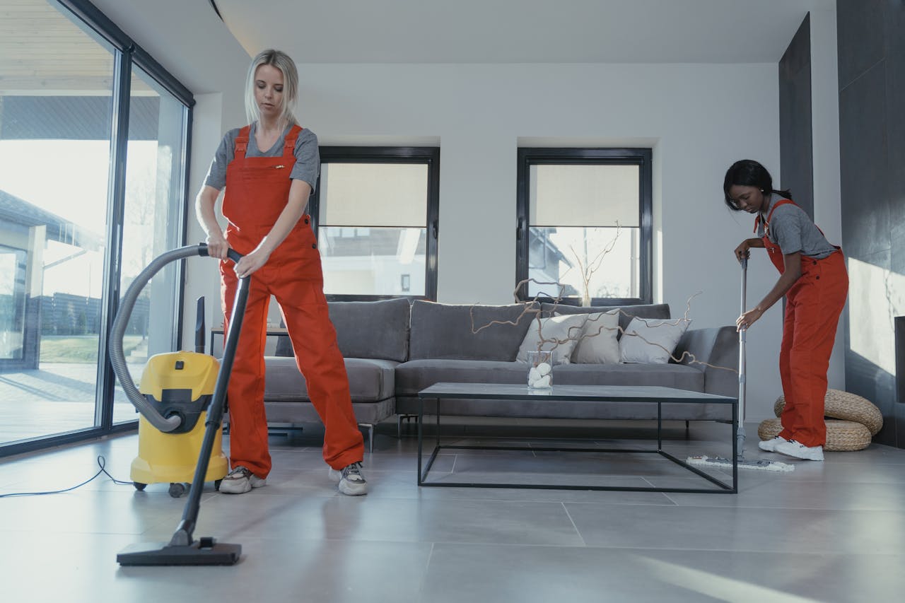 services-04 Two women in red overalls vacuuming and sweeping a modern living room.