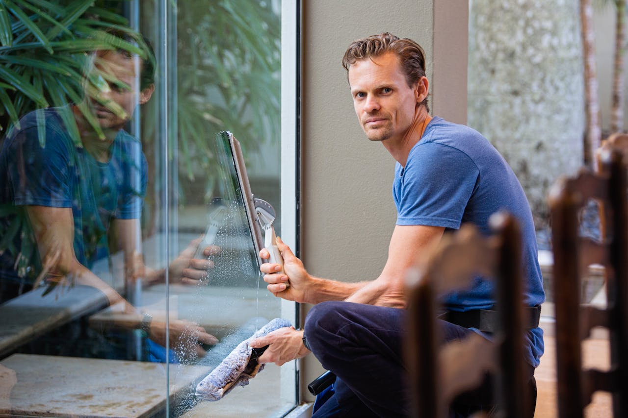 heros-img A man in a blue t-shirt cleaning large glass pane with a squeegee indoors.